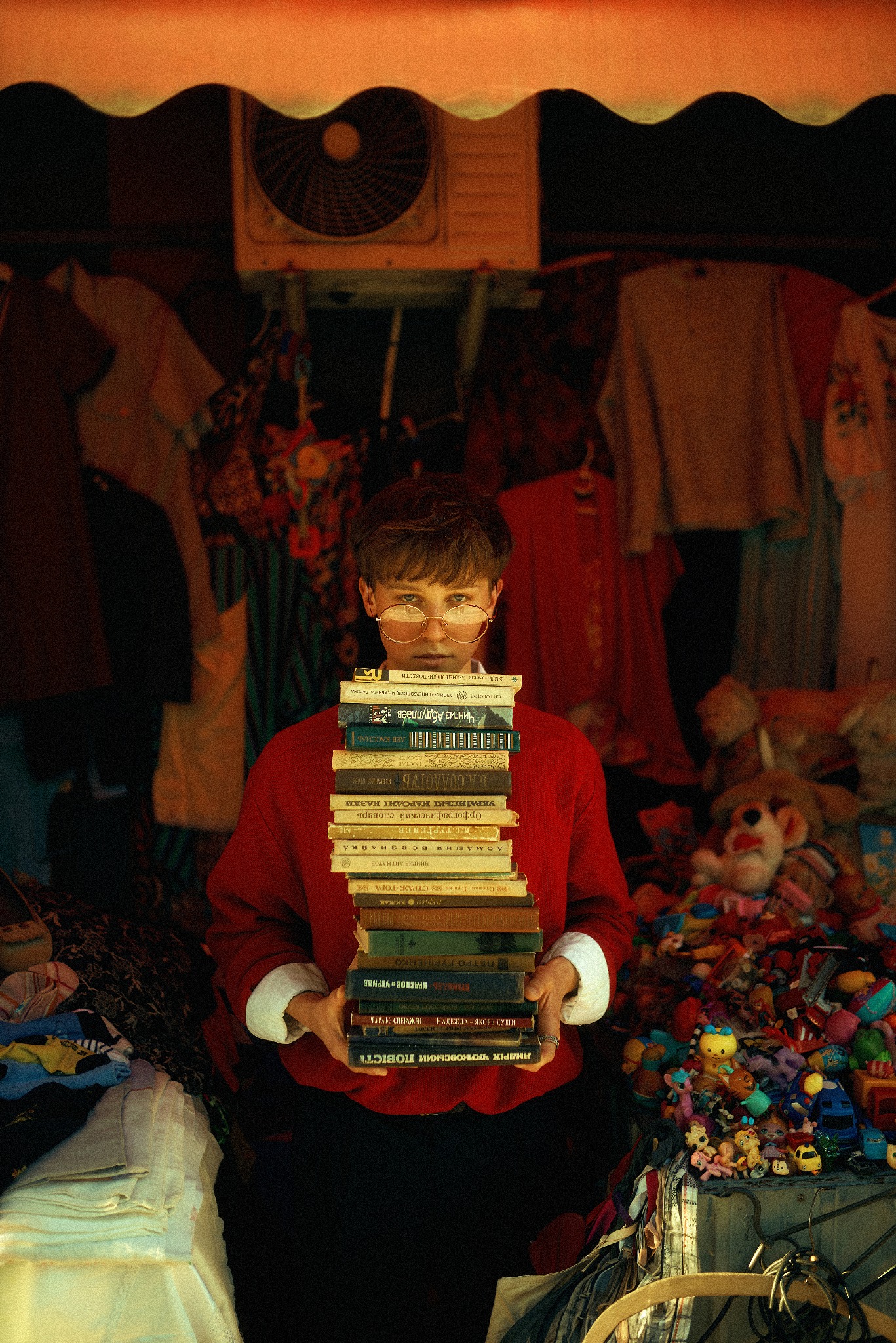 Osoba w okularach trzyma stertę książek z cyrylicą na tle straganu z ubraniami i zabawkami, oświetlona ciepłym światłem.