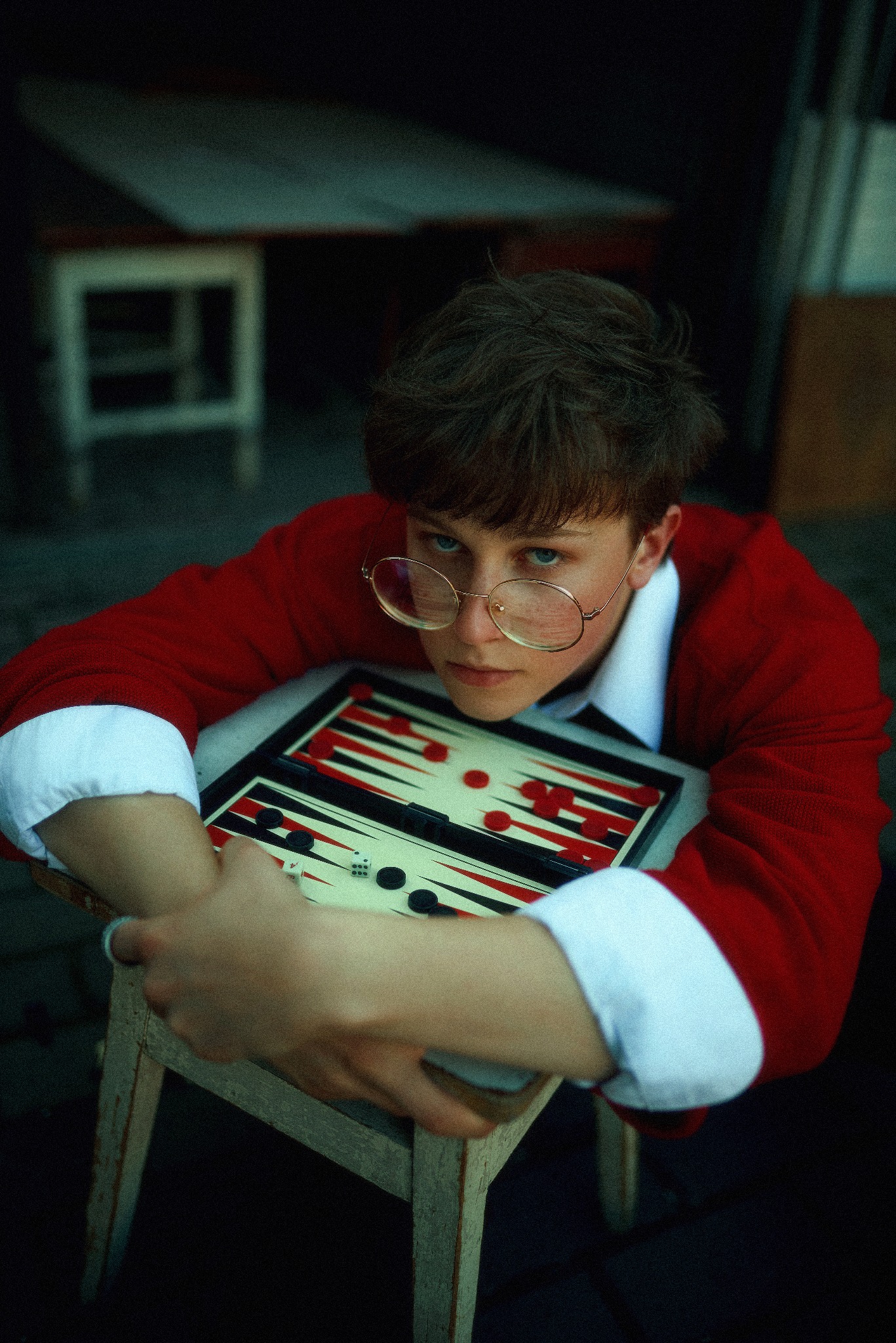 Portret młodego mężczyzny w okularach, opierającego się na stole do gry w tryktraka, z kostkami i pionkami, w czerwonym swetrze z białymi mankietami.