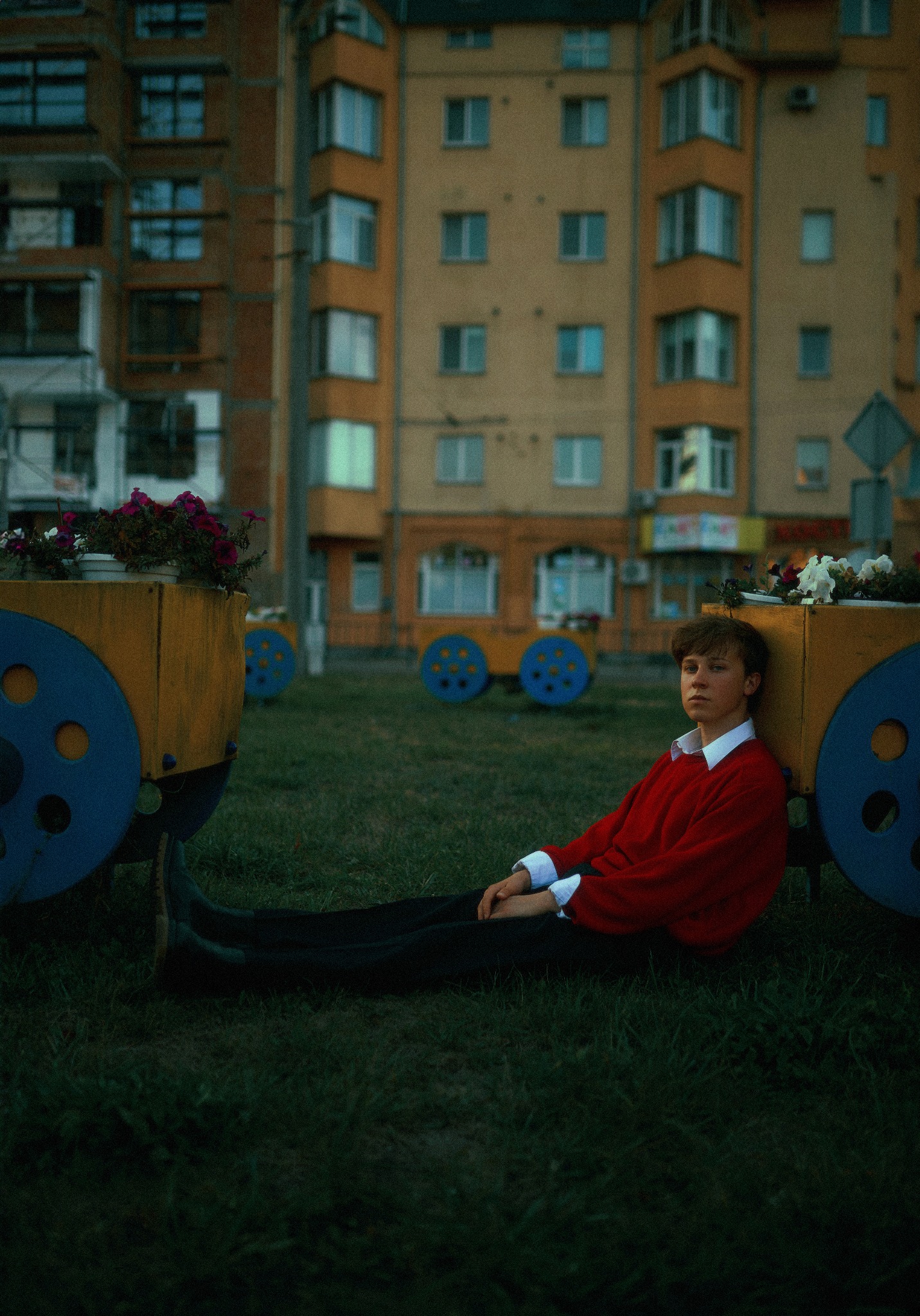 Młody mężczyzna w czerwonym swetrze oparty o dekoracyjny wózek z kwiatami na tle bloku mieszkalnego.