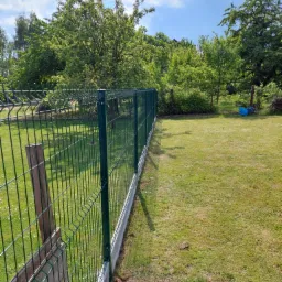 Zielone ogrodzenie panelowe z podmurówką betonową, widok z boku na tle trawnika i drzew w słoneczny dzień.