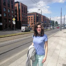 Kobieta w niebieskiej koszulce i miętowych spodniach stoi na chodniku przy ulicy z torami tramwajowymi, w tle budynki z czerwonej cegły i słupy trakcji tramwajowej.