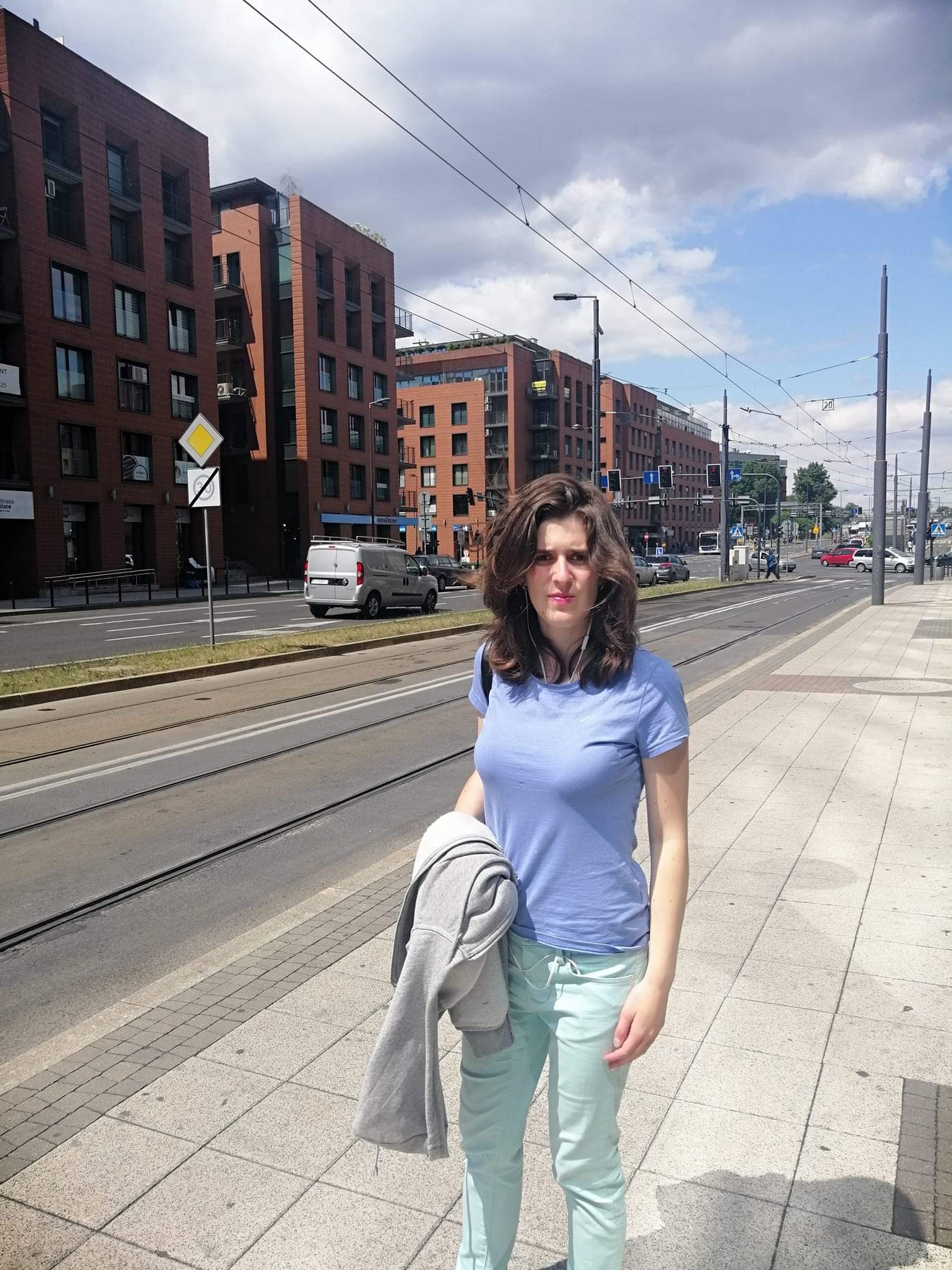 Kobieta w niebieskiej koszulce i miętowych spodniach stoi na chodniku przy ulicy z torami tramwajowymi, w tle budynki z czerwonej cegły i słupy trakcji tramwajowej.