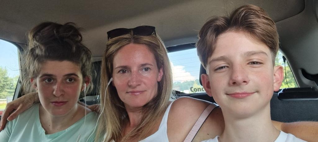 Portret matki i dwójki dzieci w samochodzie, z widokiem na budynek w tle.