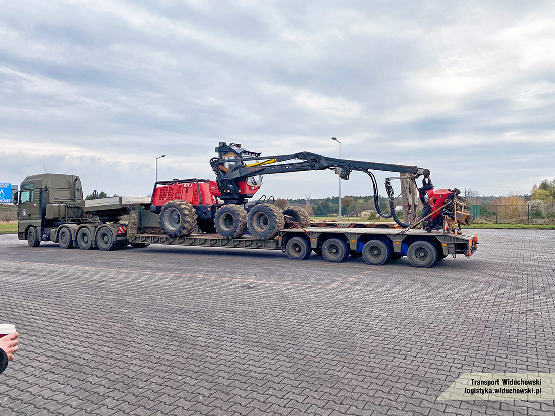 Ciężarówka z naczepą niskopodwoziową przewożąca czerwony kombajn leśny z żurawiem na placu wyłożonym kostką brukową, widoczna tablica reklamowa w tle.