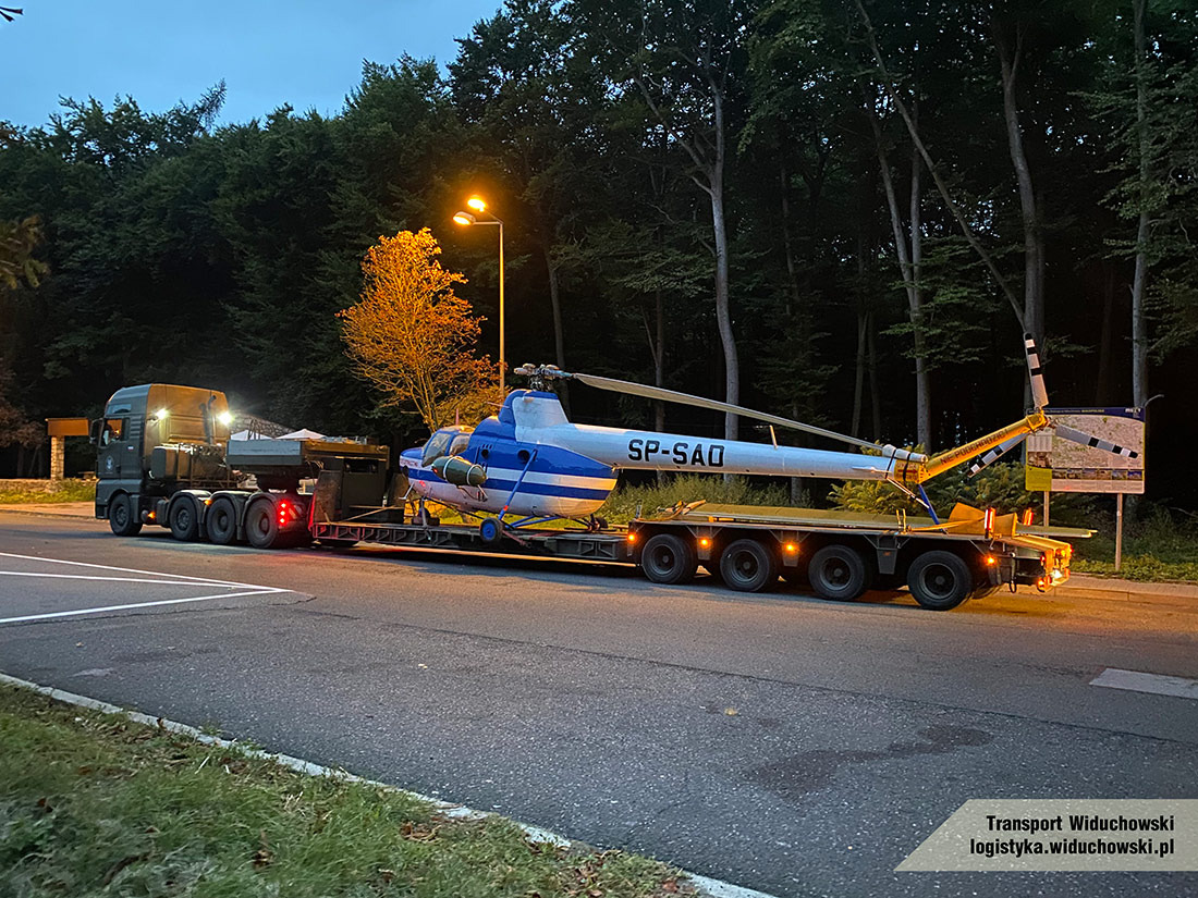 Ciężarówka z naczepą niskopodwoziową przewożąca biało-niebieski helikopter SP-SAQ na tle lasu i ulicznej latarni o zmierzchu.