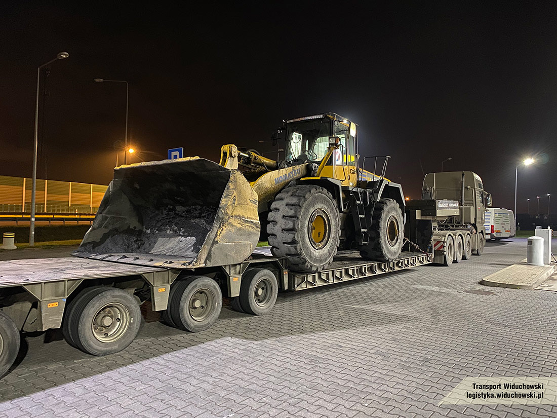 Żółta ładowarka Komatsu na niskopodwoziowej naczepie, zabezpieczona łańcuchami, przewożona nocą. Widoczny ciągnik siodłowy i fragment autobusu w tle.