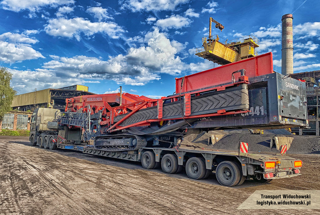 Ciężarówka transportująca na niskopodwoziowej platformie mobilny przesiewacz Sandvik Doublescreen, widoczny komin przemysłowy w tle pod błękitnym niebem z chmurami.