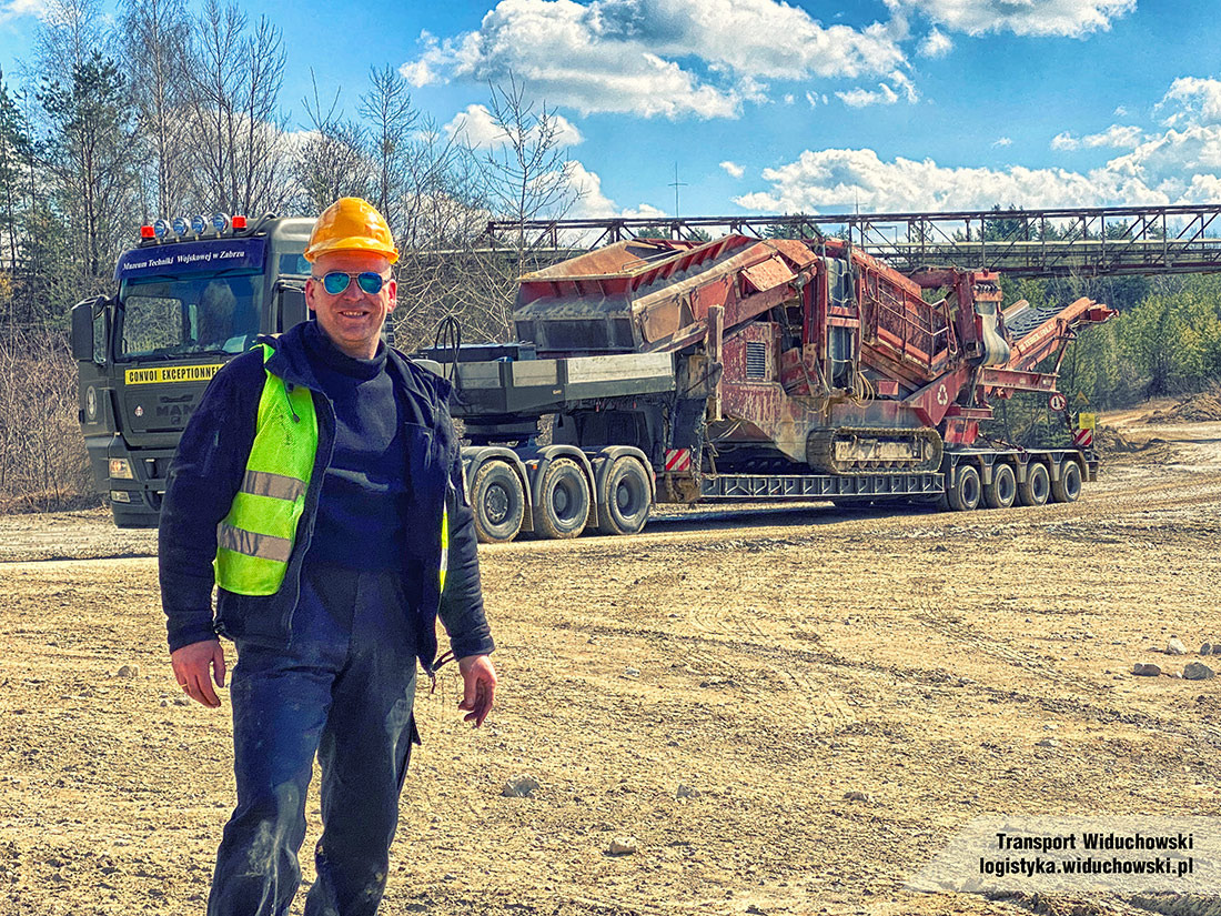 Uśmiechnięty mężczyzna w kasku i kamizelce odblaskowej stoi na tle ciężarówki z naczepą przewożącą maszynę do kruszenia skał na terenie kopalni.