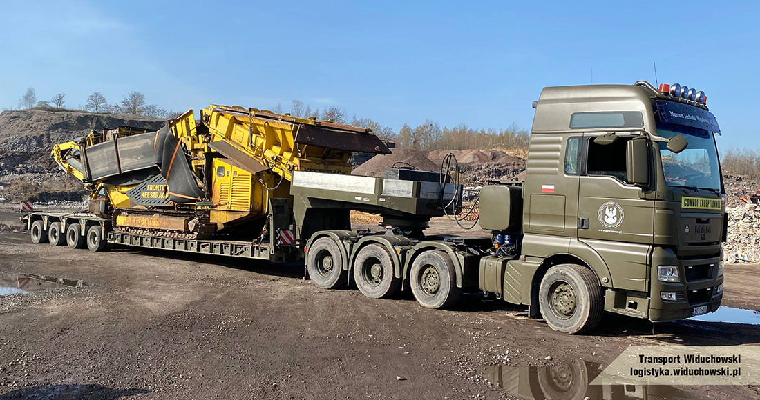 Ciężarówka MAN w kolorze khaki z polską flagą przewozi na lawecie żółtą kruszarkę do kamienia marki Fronti Keestrack na tle kamieniołomu.
