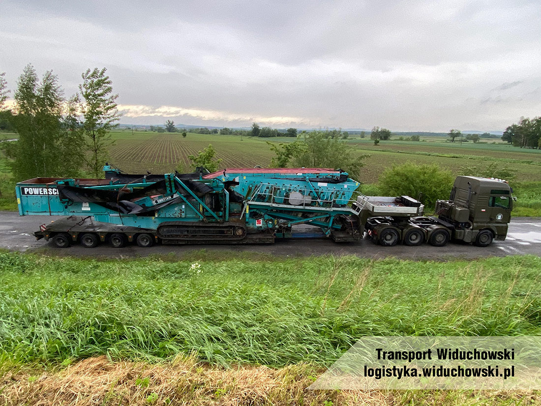 Ciężarówka transportuje mobilną kruszarkę do kamienia na platformie, widok z boku na tle zielonych pól i pochmurnego nieba.