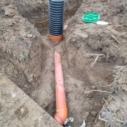 AmelBud Michał Wodecki - Wykop w ziemi z ułożoną pomarańczową rurą kanalizacyjną i czarną rurą karbowaną stojącą pionowo; zielona pokrywa studzienki obok.