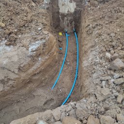 AmelBud Michał Wodecki - Wykop w ziemi z widocznymi dwiema niebieskimi rurami, z fragmentami śniegu w tle i betonową studzienką kanalizacyjną.