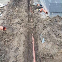 AmelBud Michał Wodecki - Wykopany rów z ułożonymi pomarańczowymi rurami kanalizacyjnymi, widoczny fragment budynku z szarych bloczków i koparka w tle.