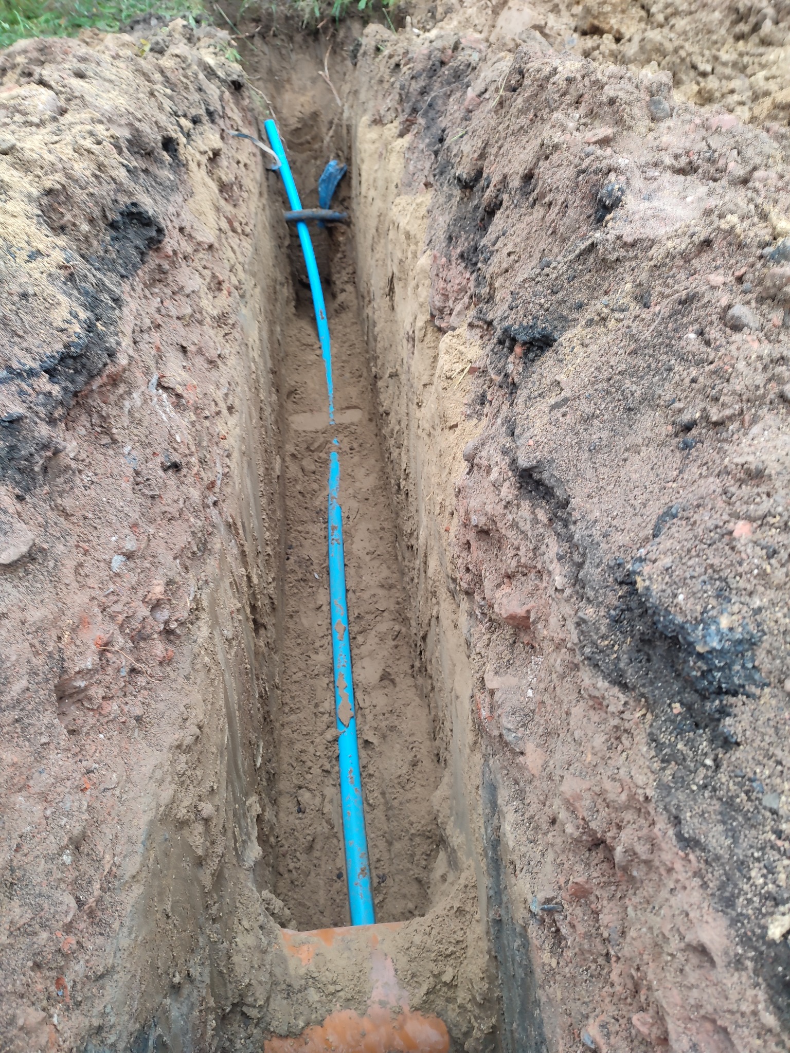Wykop w ziemi z widoczną niebieską rurą instalacyjną i pomarańczową rurą kanalizacyjną na dnie wykopu.