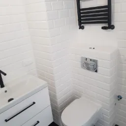 Nowoczesna łazienka z białymi, błyszczącymi płytkami typu metro, czarną armaturą i grzejnikiem drabinkowym, oraz podłogą w kolorze czarnym.
