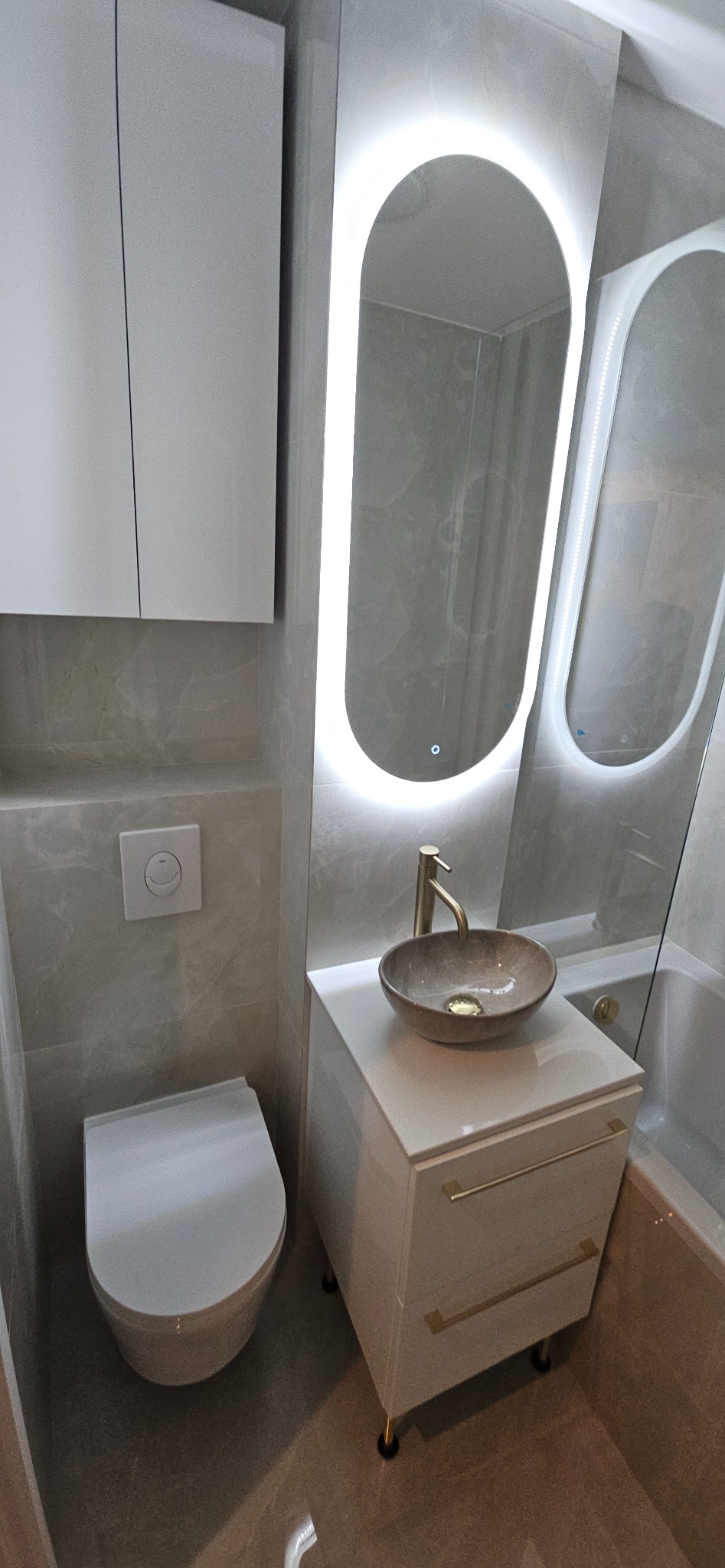 Nowoczesna łazienka z podświetlanym lustrem, umywalką nablatową i wiszącą toaletą. Jasne płytki imitujące marmur. Złote akcenty na baterii i nóżkach szafki.