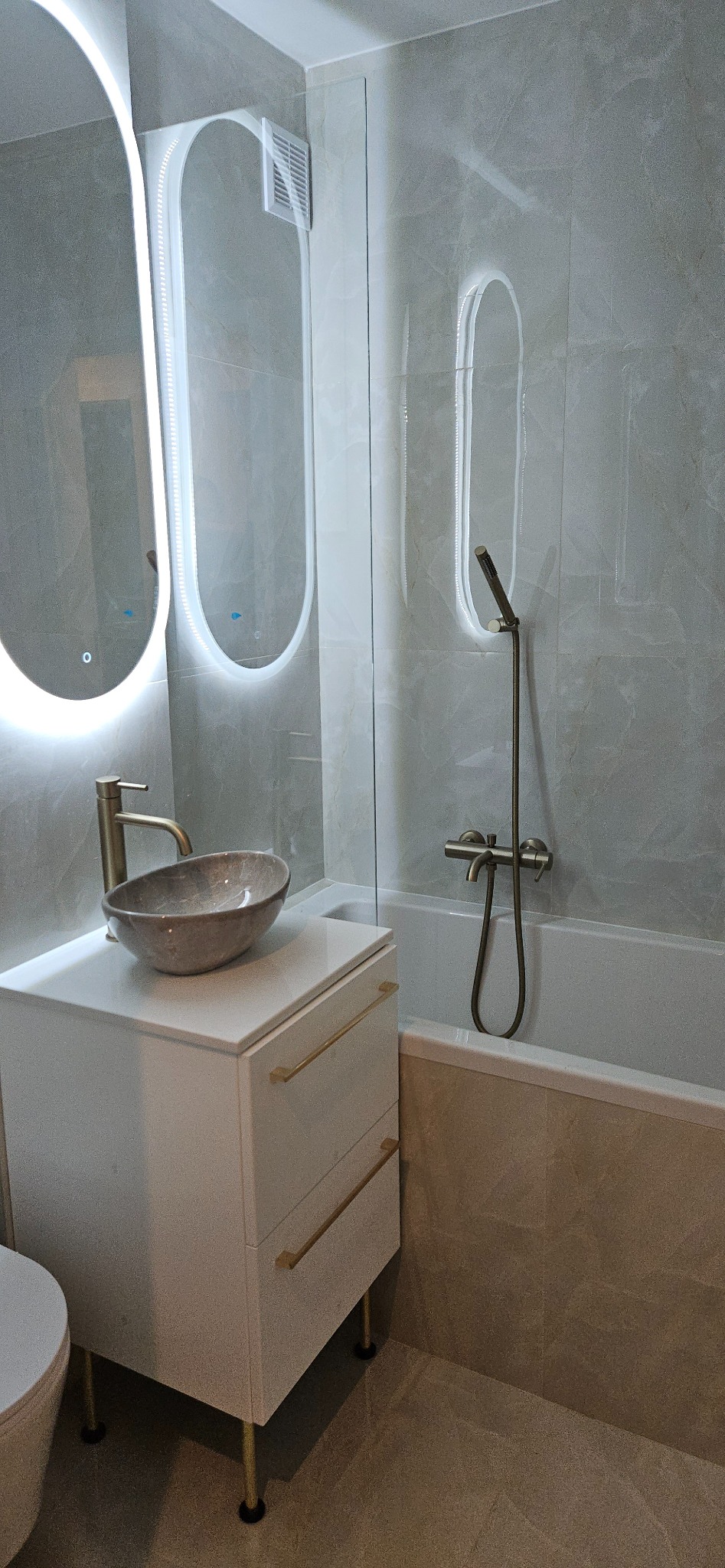 Nowoczesna łazienka z umywalką nablatową, podświetlanym lustrem i szklanym parawanem nawannowym. Minimalistyczny design, szare płytki i złote akcenty.