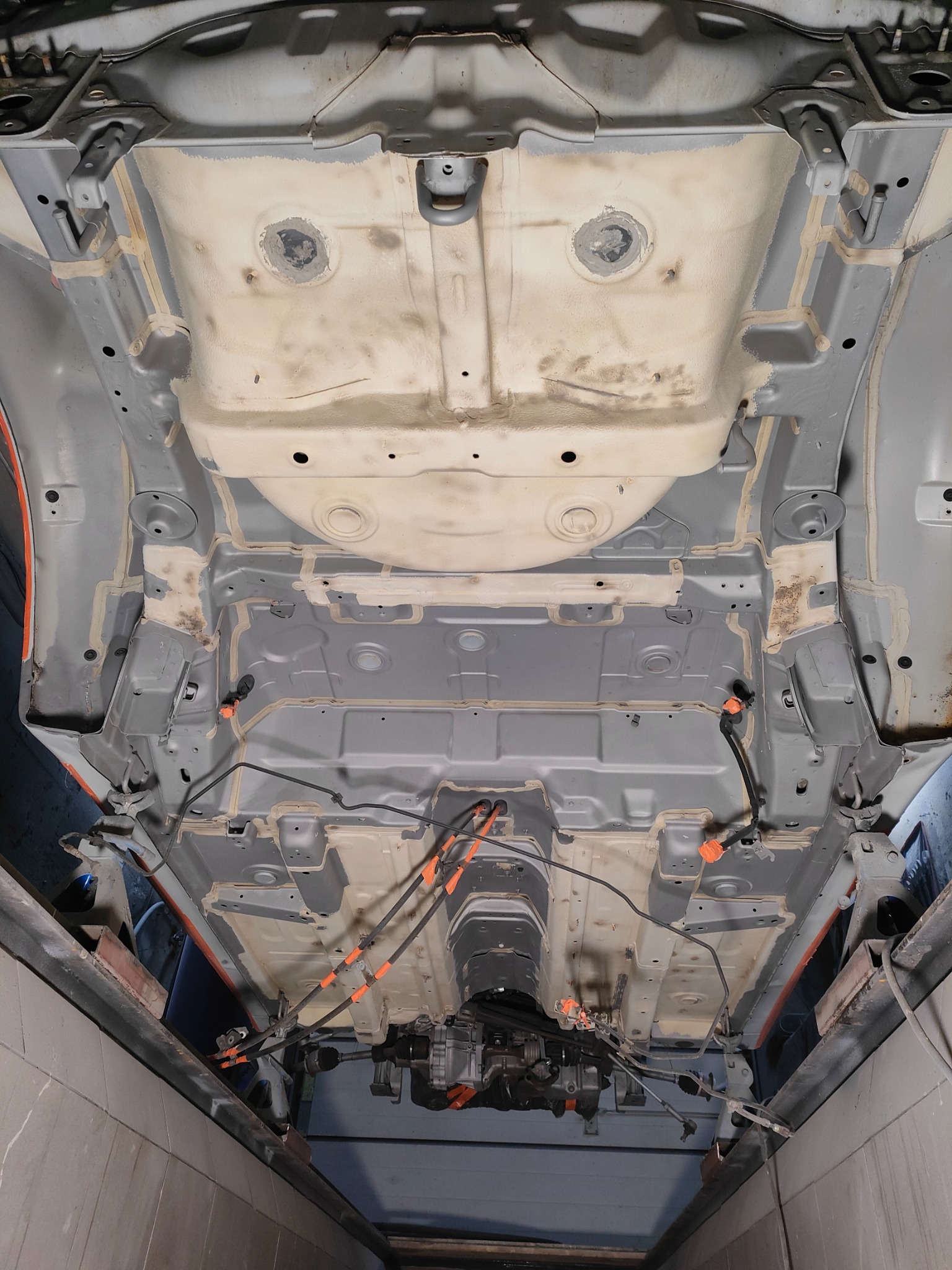 Podwozie samochodu po zabezpieczeniu antykorozyjnym, widoczne elementy konstrukcyjne i zabezpieczenia, zarys podnośnika warsztatowego w tle.