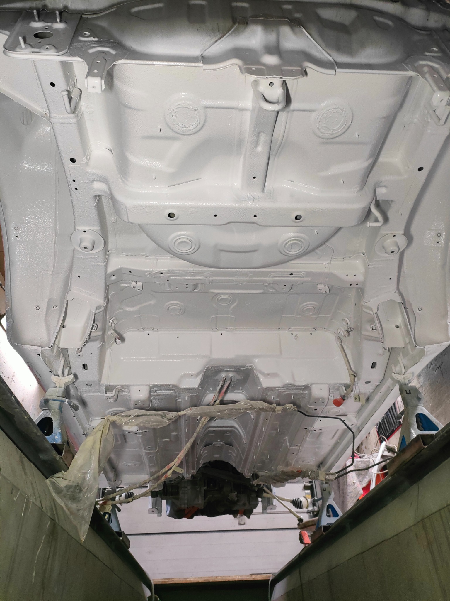 Spód samochodu po zabezpieczeniu antykorozyjnym i pomalowaniu na biało, widoczne elementy zawieszenia, podnośniki warsztatowe i folia ochronna.