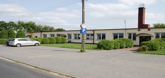 Budynek szwalni z parkującymi samochodami, widoczny znak parkingu, krzewy ozdobne i komin wentylacyjny.