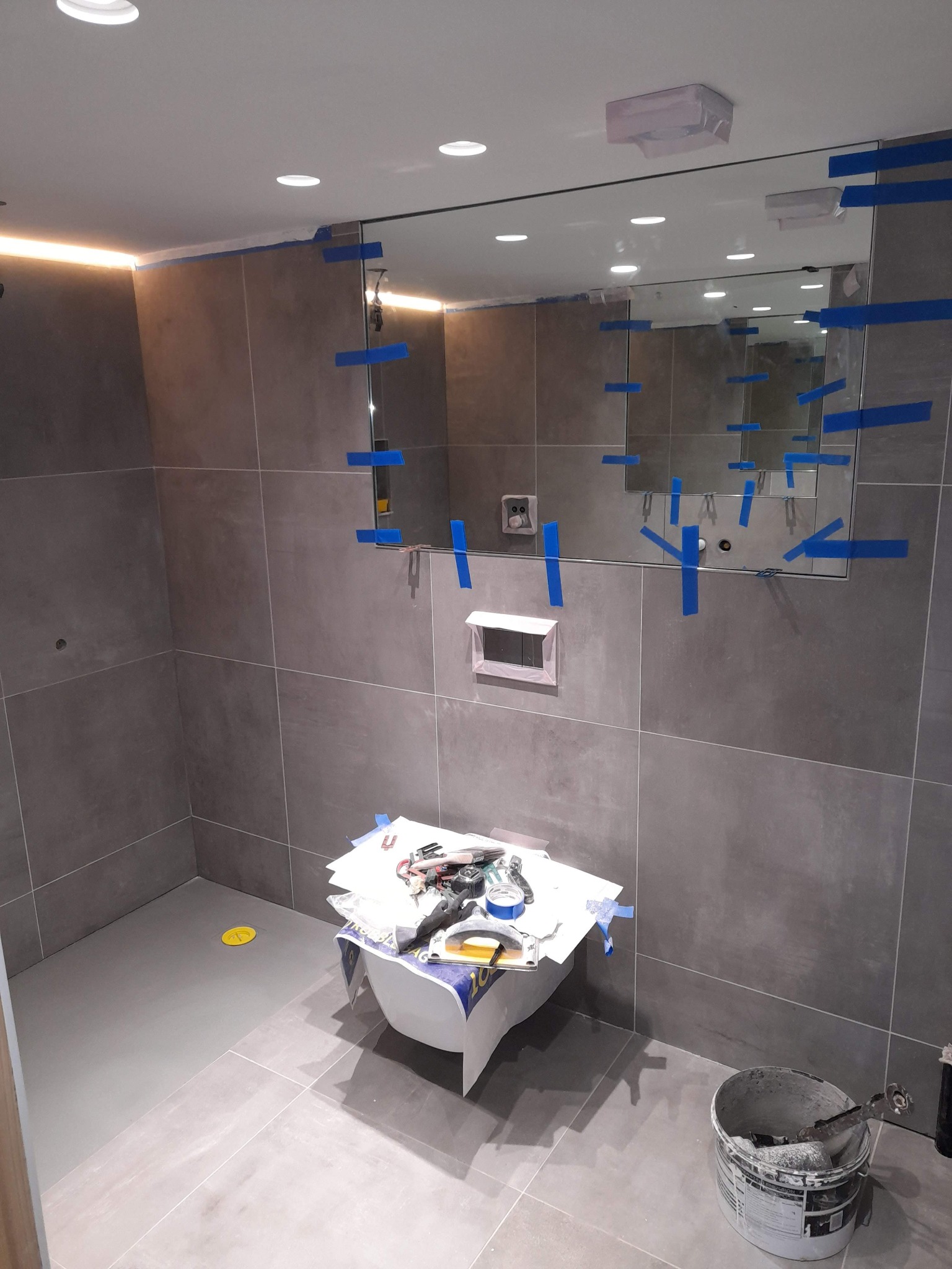 Łazienka w trakcie prac z szarymi płytkami, zabezpieczone lustro niebieską taśmą, widoczne narzędzia i wiadro z zaprawą.