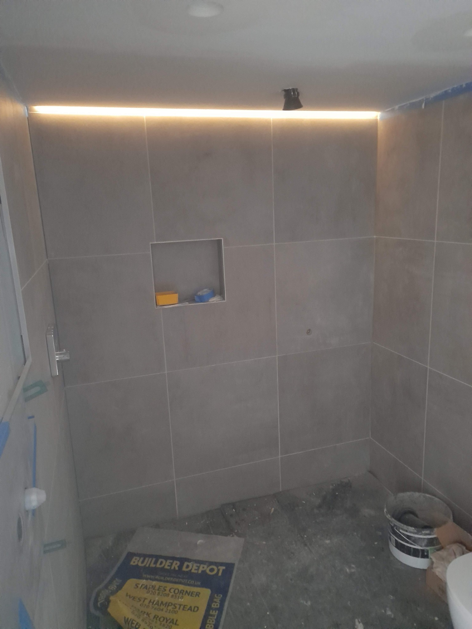 Szara łazienka w trakcie wykańczania, z podświetleniem LED podsufitowym, wnęką w ścianie, widoczne materiały budowlane i wiadro na podłodze.