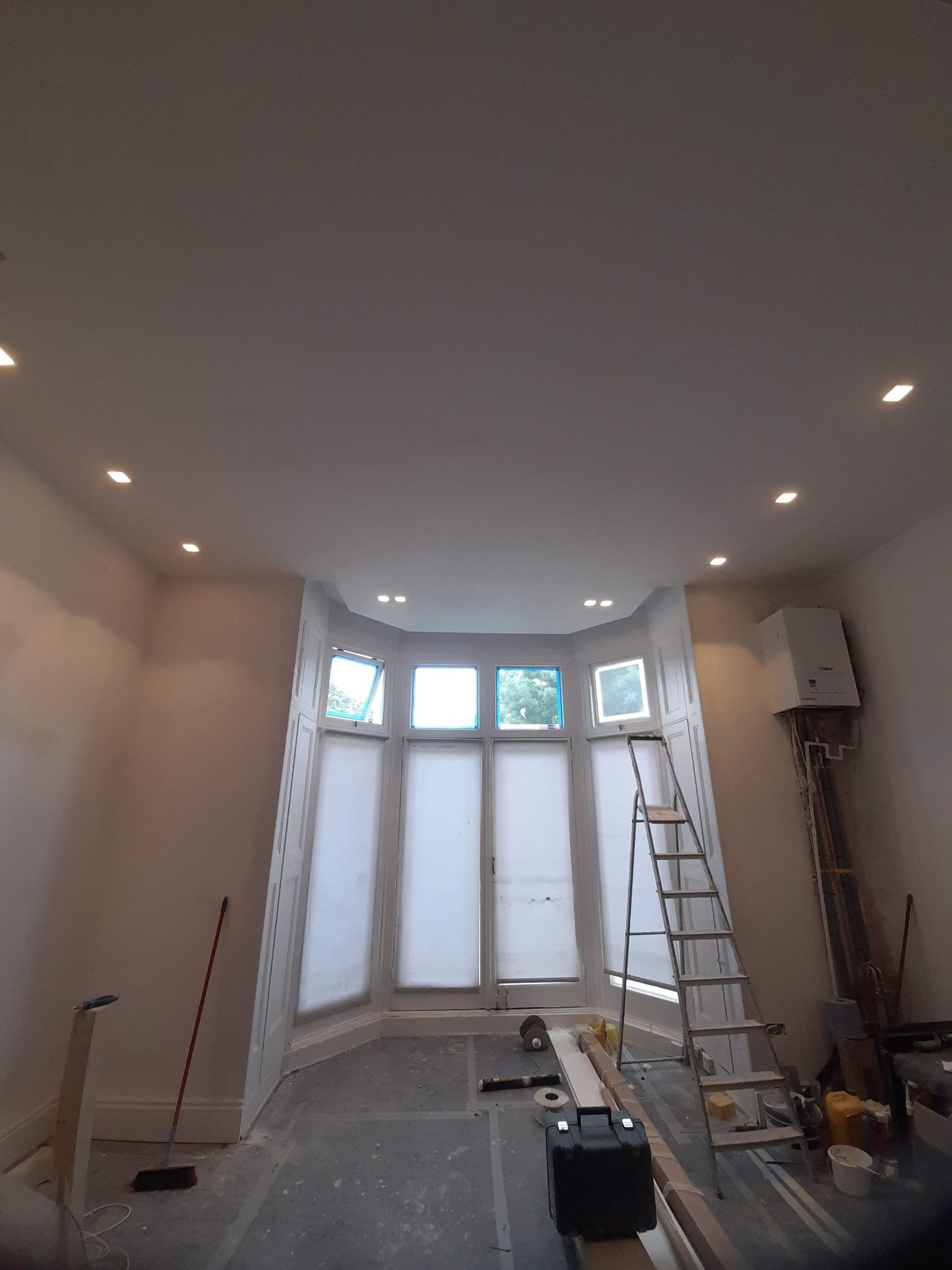 Pokój w trakcie prac, widoczne okna z osłonami, drabina, narzędzia, świeżo pomalowane ściany z zamontowanym oświetleniem punktowym oraz piec gazowy na ścianie.