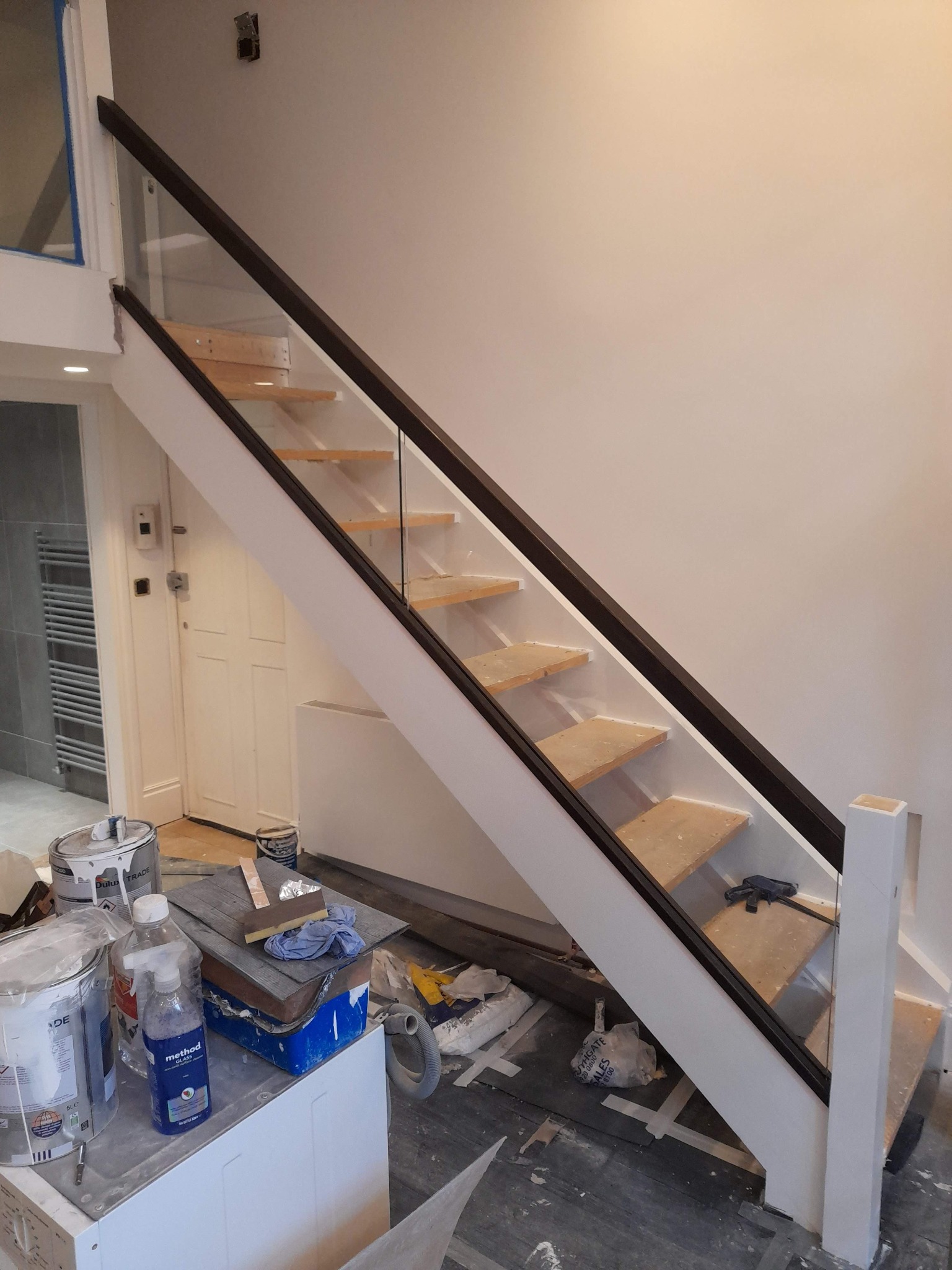 Nowoczesne schody z drewnianymi stopniami i szklaną balustradą w trakcie montażu, widoczne narzędzia i materiały malarskie, wnętrze w trakcie wykończenia.