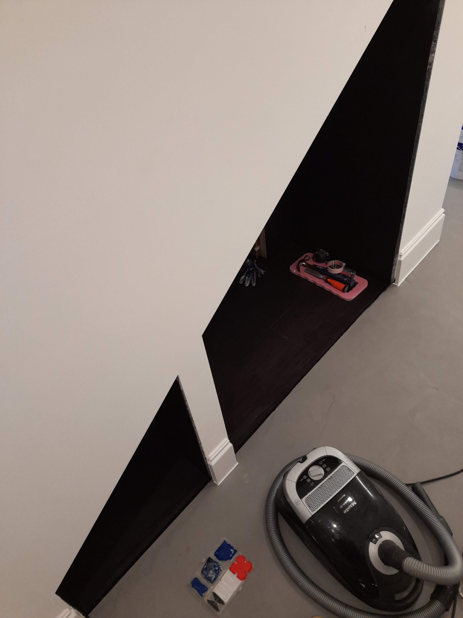 Wnęka pod schodami pomalowana na czarno, z narzędziami na różowej tacy wewnątrz, obok odkurzacz Miele i opakowania po materiałach budowlanych.