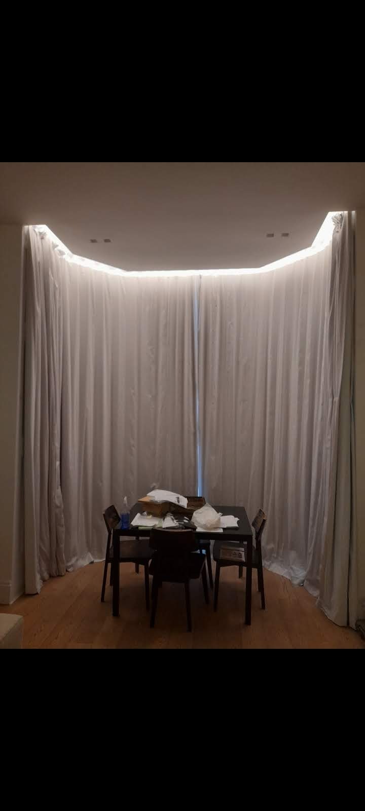 Pokój z drewnianą podłogą, czarnym stołem i krzesłami otoczony jasnymi, zwisającymi zasłonami z oświetleniem LED ukrytym w suficie.