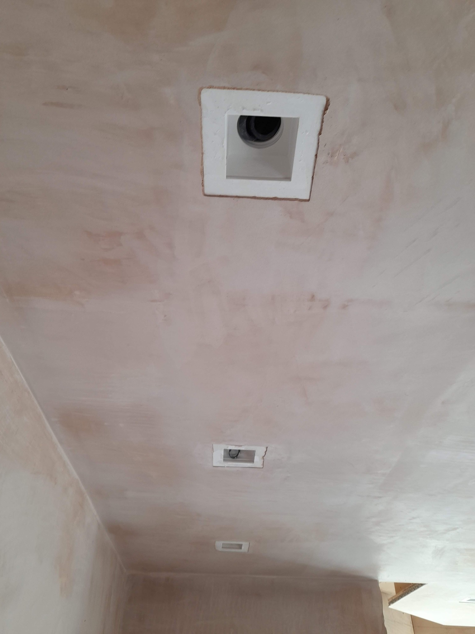 Niedokończona powierzchnia sufitu z widocznymi śladami szpachlowania i dwoma kwadratowymi otworami wentylacyjnymi, jeden z kratką, drugi z czarnym wlotem do kanału wentylacyjnego.
