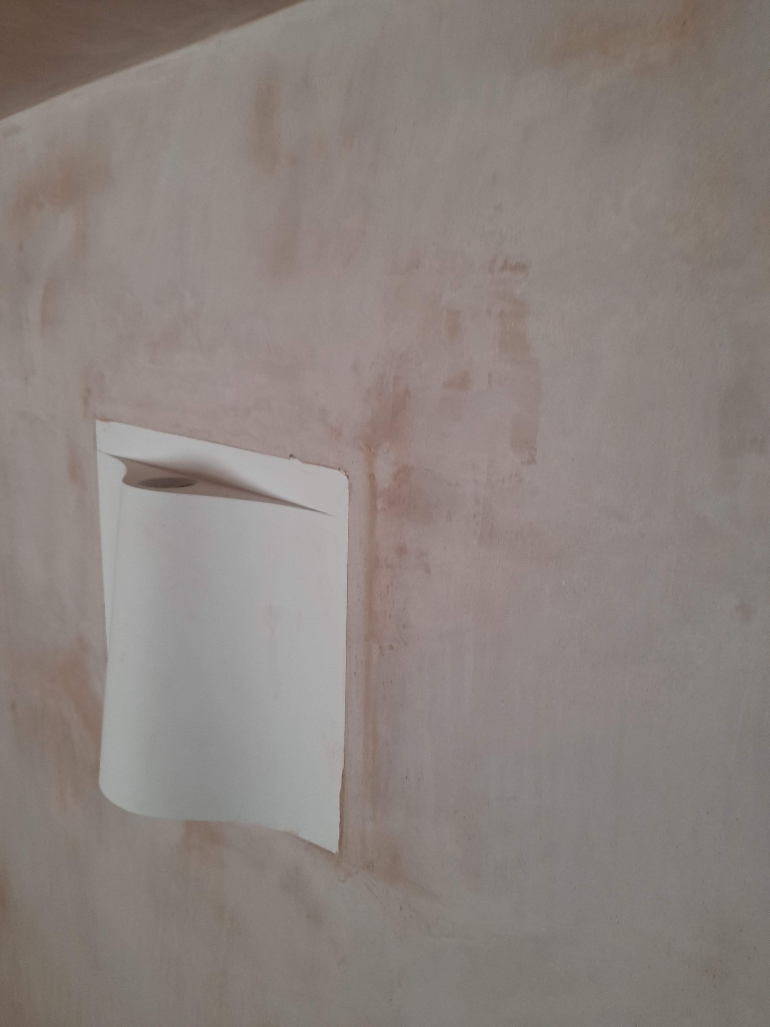 Nierówna, jasna ściana w trakcie prac, z fragmentem przyklejonej taśmy malarskiej odrywającej się w górnym rogu.