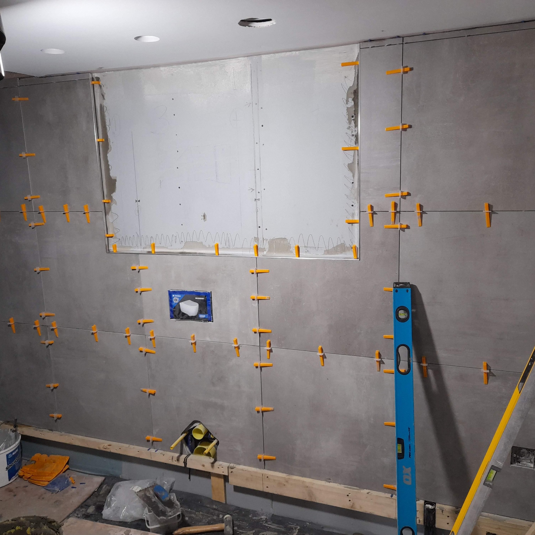 Układanie szarych płytek na ścianie z użyciem poziomicy i pomarańczowych krzyżyków dystansowych, widoczna puszka z logo Geberit