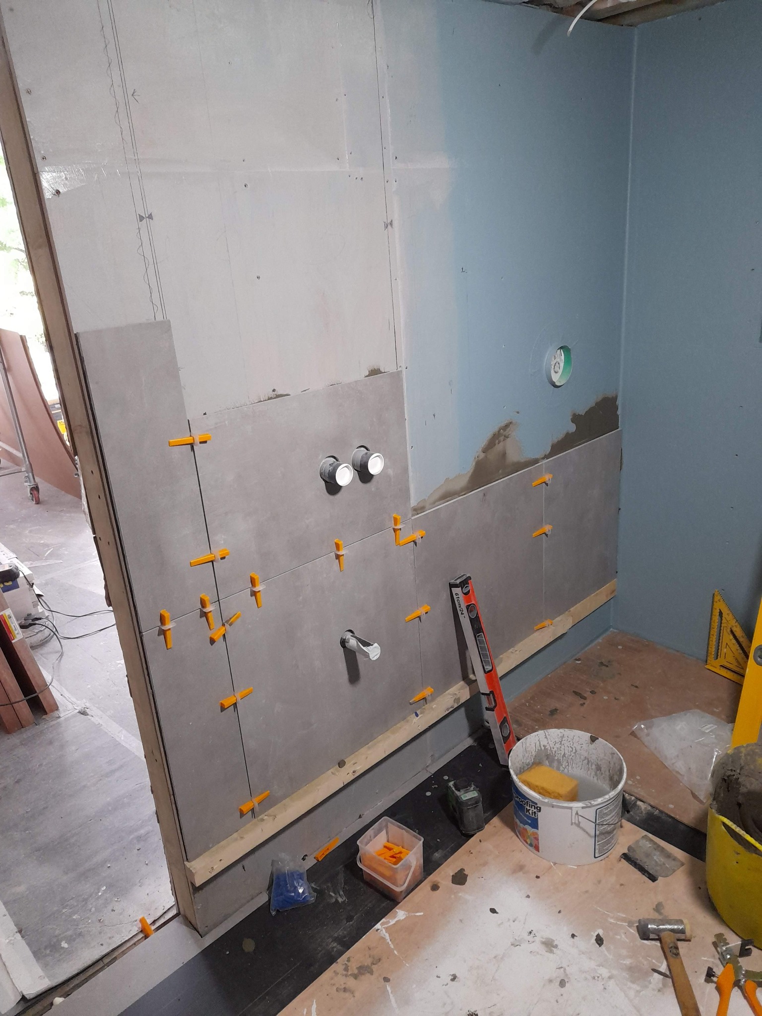 Montaż płytek ceramicznych na ścianie w łazience, widoczne elementy instalacji hydraulicznej, poziomica i krzyżyki dystansowe, na podłodze narzędzia i wiadro z zaprawą.