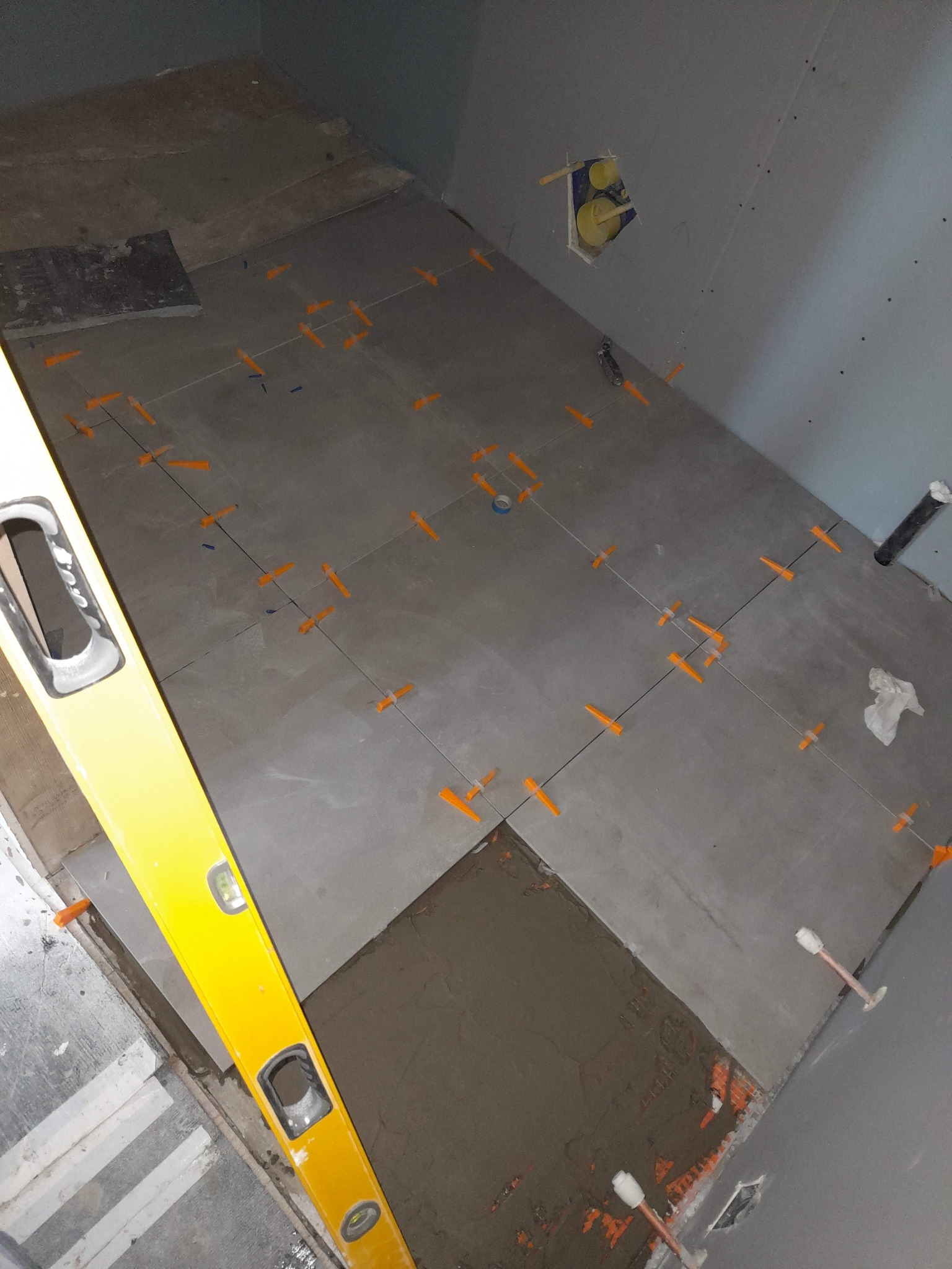 Układanie szarych płytek podłogowych z użyciem pomarańczowych klinów poziomujących, widoczna poziomica i fragment nałożonego kleju.