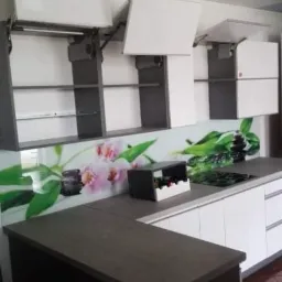 Nowoczesna kuchnia z białymi frontami szafek otwieranych do góry, szarymi korpusami i blatem w kolorze ciemnego drewna, ozdobiona panelem szklanym z motywem kwiatowym.