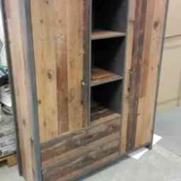 Rustykalna szafa z drewnianymi frontami z odzysku i metalową ramą, widoczne drzwi, szuflady i półki, w tle kartony.