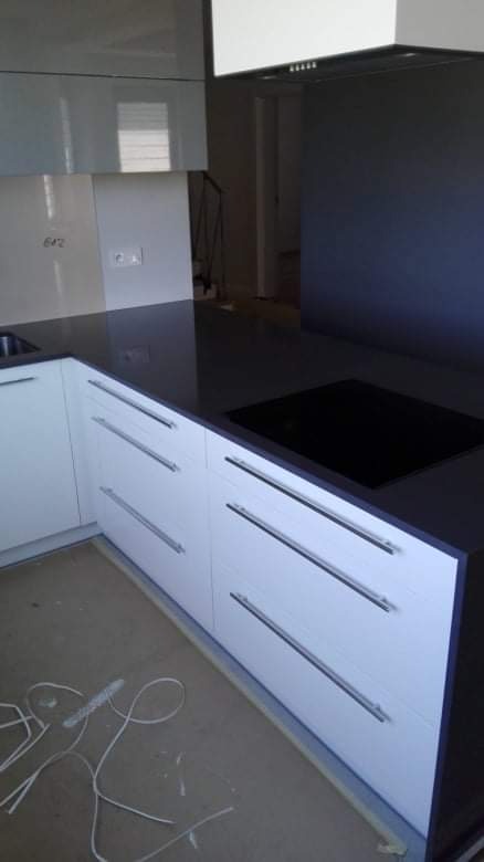 Nowoczesna kuchnia z białymi szafkami z długimi, poziomymi uchwytami i czarnym blatem, na którym zamontowana jest płyta indukcyjna. Na podłodze leżą resztki taśmy malarskiej.