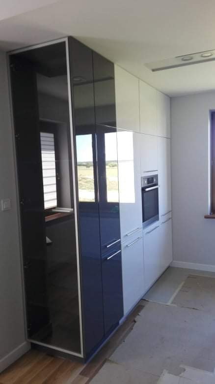 Nowoczesna zabudowa kuchenna z wysokimi szafkami w kolorze białym i granatowym z połyskiem, z wbudowanym piekarnikiem, widok na okno z roletami i krajobraz za nim, podłoga w trakcie prac...