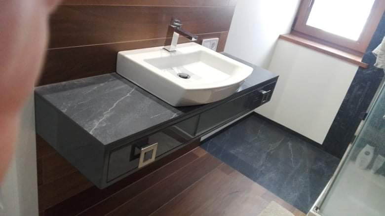 Nowoczesna łazienka z umywalką nablatową na ciemnoszarym blacie z szufladą, drewniana boazeria na ścianie, widoczny fragment kabiny prysznicowej.