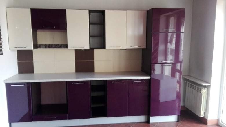Nowoczesna zabudowa kuchenna w odcieniach fioletu i beżu, z błyszczącymi frontami i płytkami w pasującym kolorze, widoczny grzejnik pod oknem.