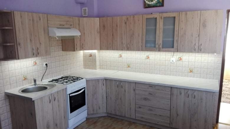 Jasna kuchnia w zabudowie z drewnopodobnymi frontami, widoczny zlew, płyta gazowa i piekarnik, ściana wyłożona kwadratowymi płytkami z delikatnym wzorem, oświetlenie podszafkowe.