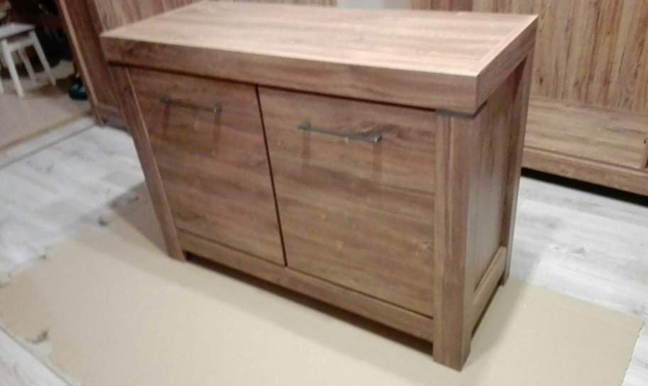 Dwudrzwiowa komoda z drewna o ciepłym odcieniu, z prostymi, metalowymi uchwytami, ustawiona na tekturowej podkładce w pomieszczeniu z drewnianą podłogą i widocznymi innymi meblami w tle.