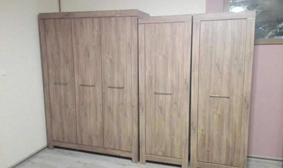 Komplet szaf z drewnopodobnymi frontami, z podłużnymi uchwytami, ustawiony w pomieszczeniu z widoczną ścianą w kolorze pudrowego różu.