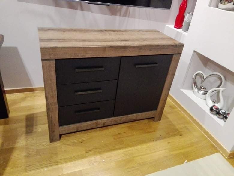 Komoda z drewnianym blatem i czarnymi frontami szuflad i szafki, ustawiona na parkiecie, z dekoracjami w tle.