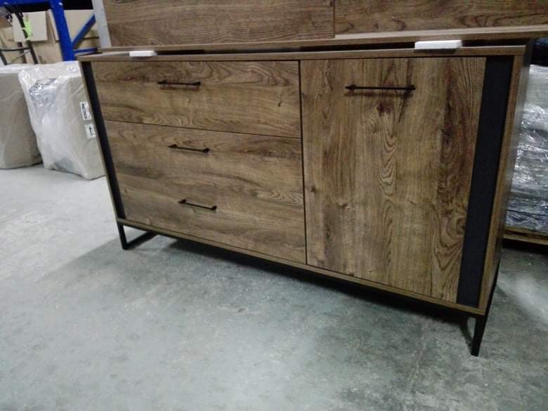 Komoda z trzema szufladami i szafką, okleina w kolorze drewna, czarne metalowe nóżki i uchwyty, widoczna w magazynie.