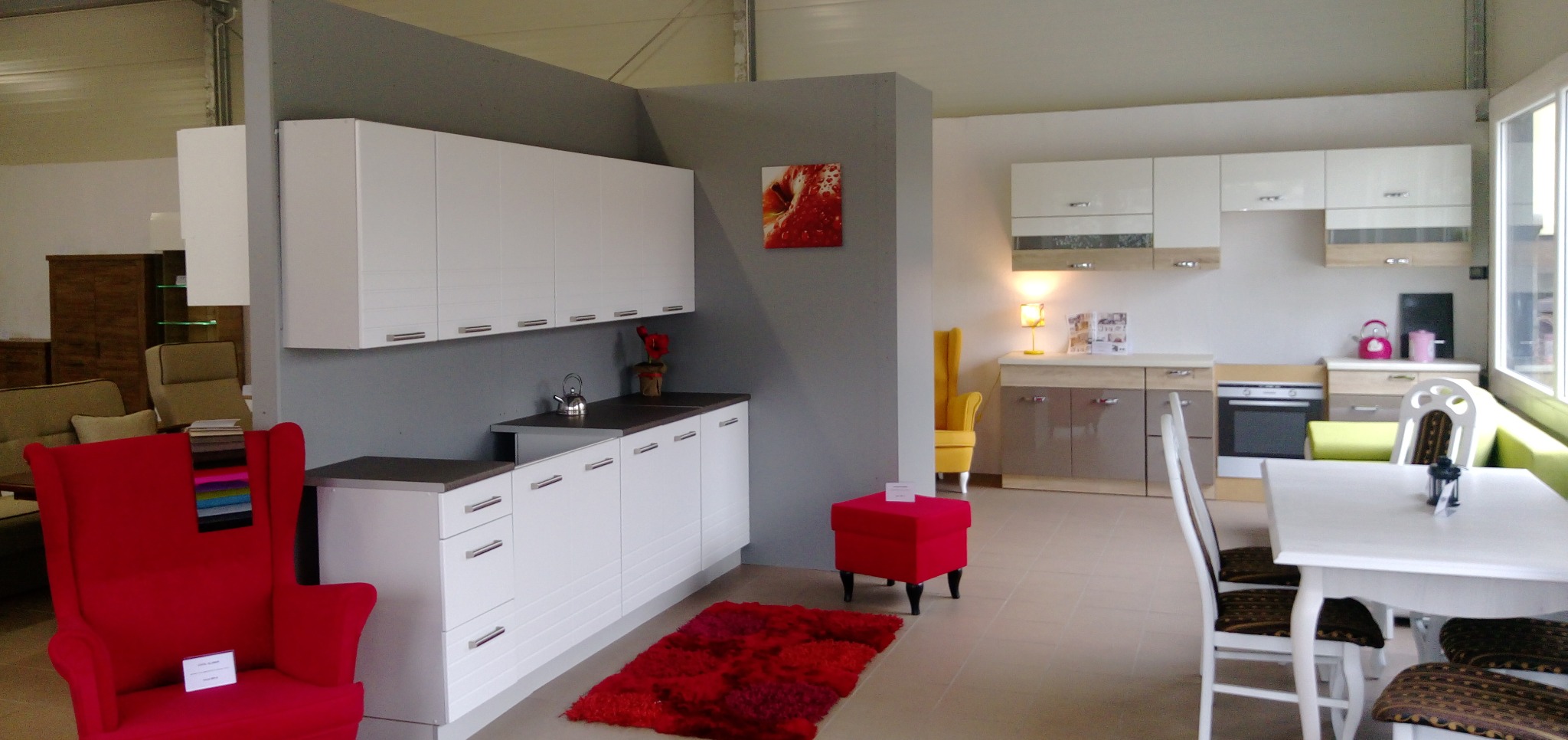 Wnętrze ekspozycji meblowej: białe szafki kuchenne z szarymi blatami, czerwony dywanik i fotel z próbkami tkanin, w tle żółty fotel i biały stół z krzesłami.