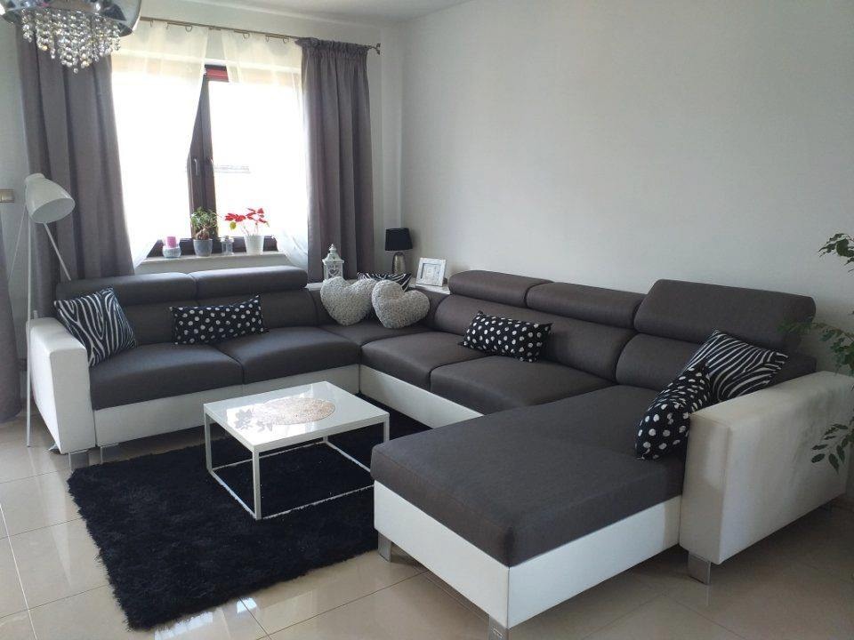 Szary narożnik z białymi bokami, wzorzyste poduszki, biały stolik kawowy na czarnym dywanie, widok na okno z szarymi zasłonami i roślinami.