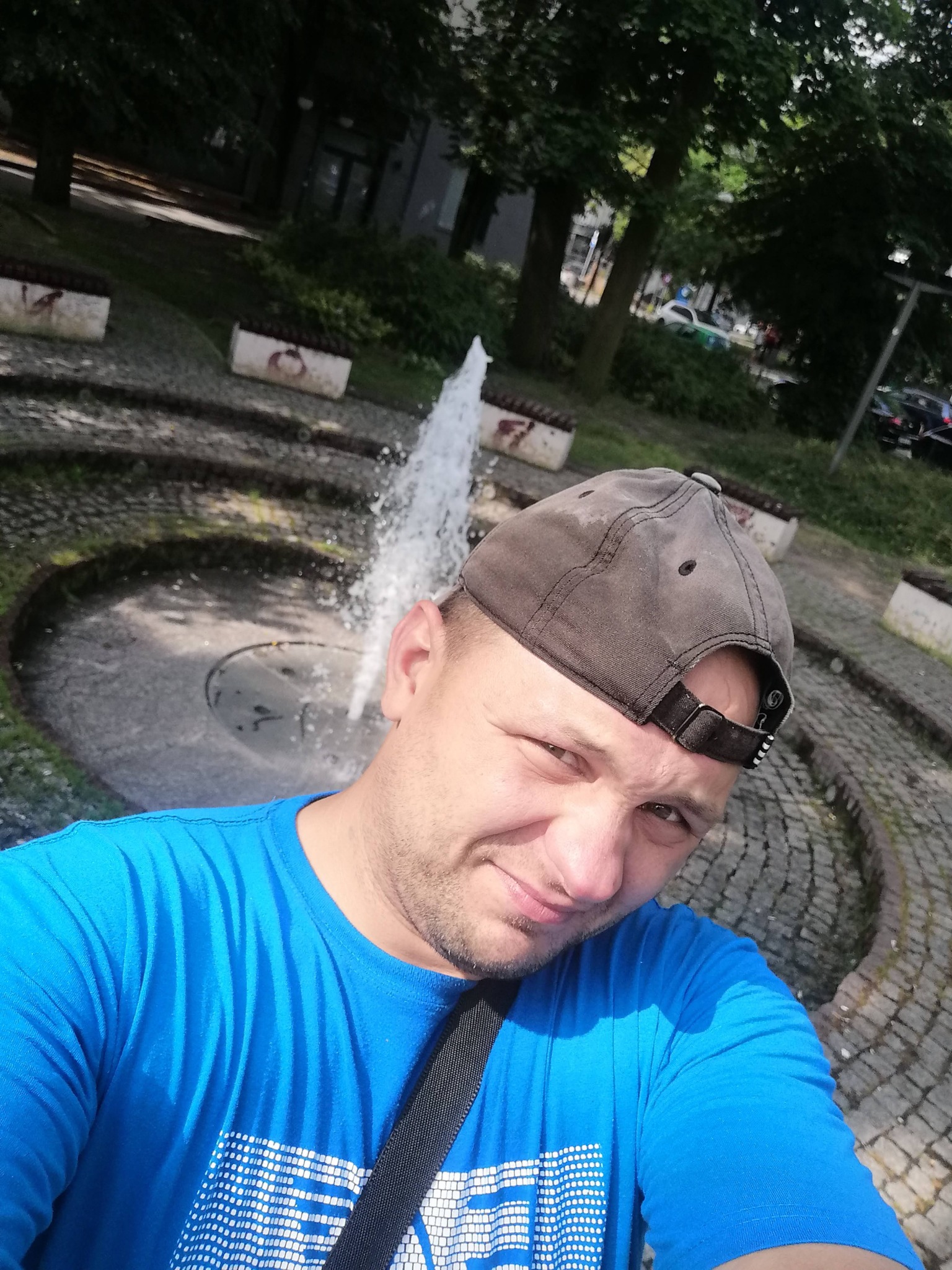 Mężczyzna w niebieskiej koszulce i czapce pozuje na tle fontanny w parku, z widocznymi drzewami i budynkami w tle.