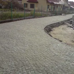 Ułożona, zakrzywiona ścieżka z kostki brukowej na podwórku, z widocznymi niedokończonymi obszarami, materiałami budowlanymi i zielonym ogrodzeniem w tle.